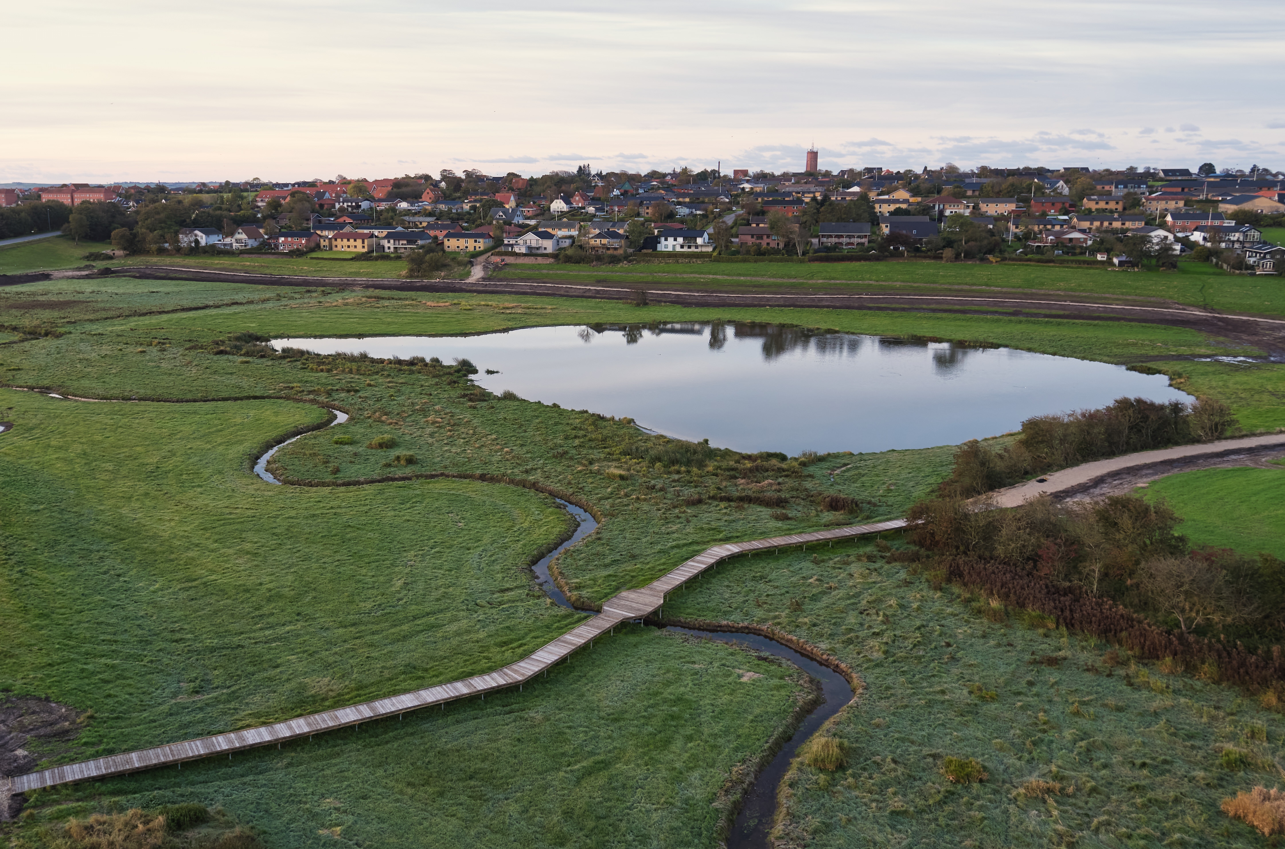 Naturplejeprojekt i et sumpet område ved en sø. En gangbro af træ snor sig gennem det våde landskab og giver adgang til området uden at forstyrre naturen. I baggrunden ses en by med farverige huse og en kirke.