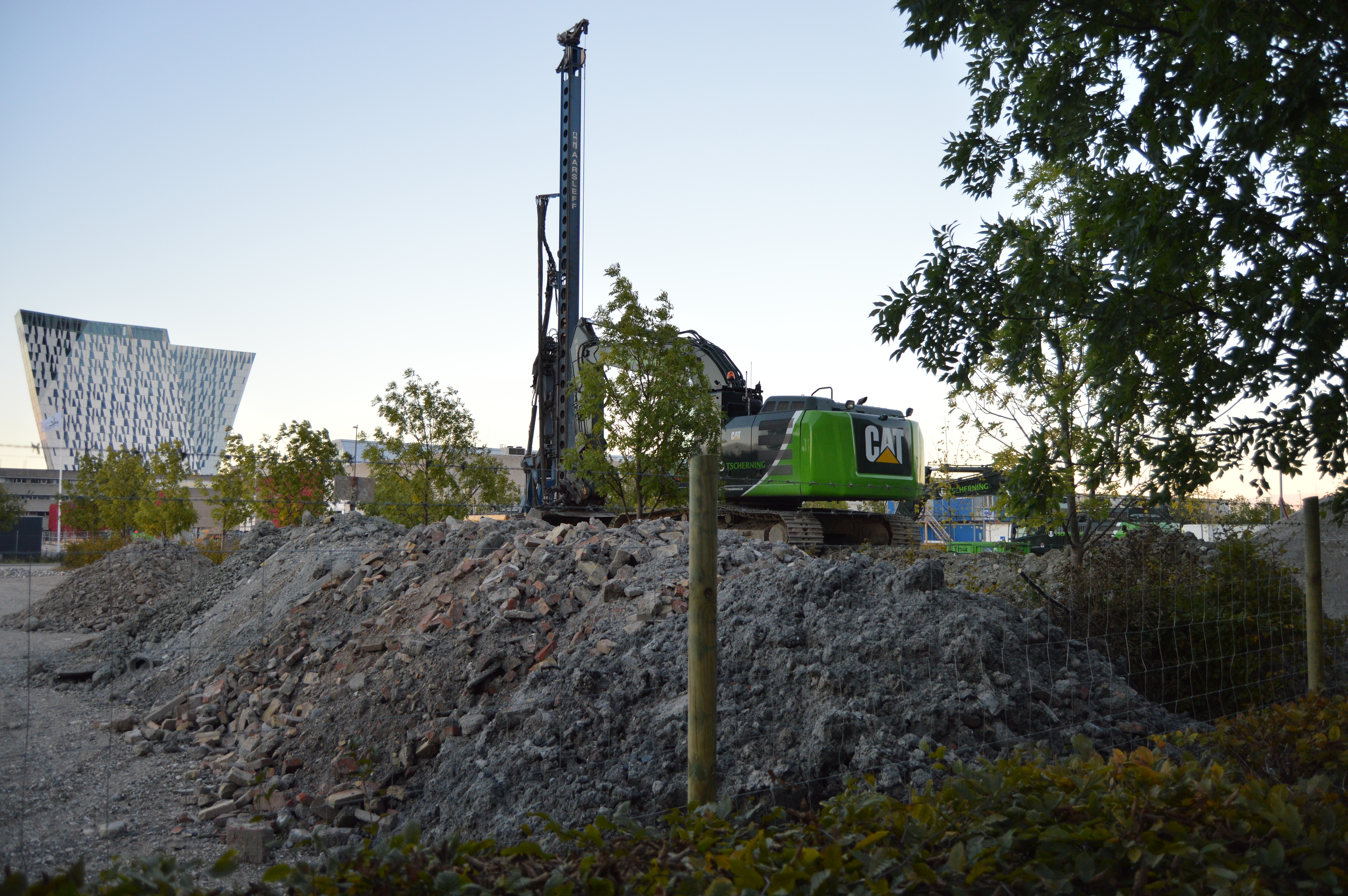 En grøn CAT maskine holder parkeret ovenpå en grusbunke. Maskinen er sikret med et byggepladshegn