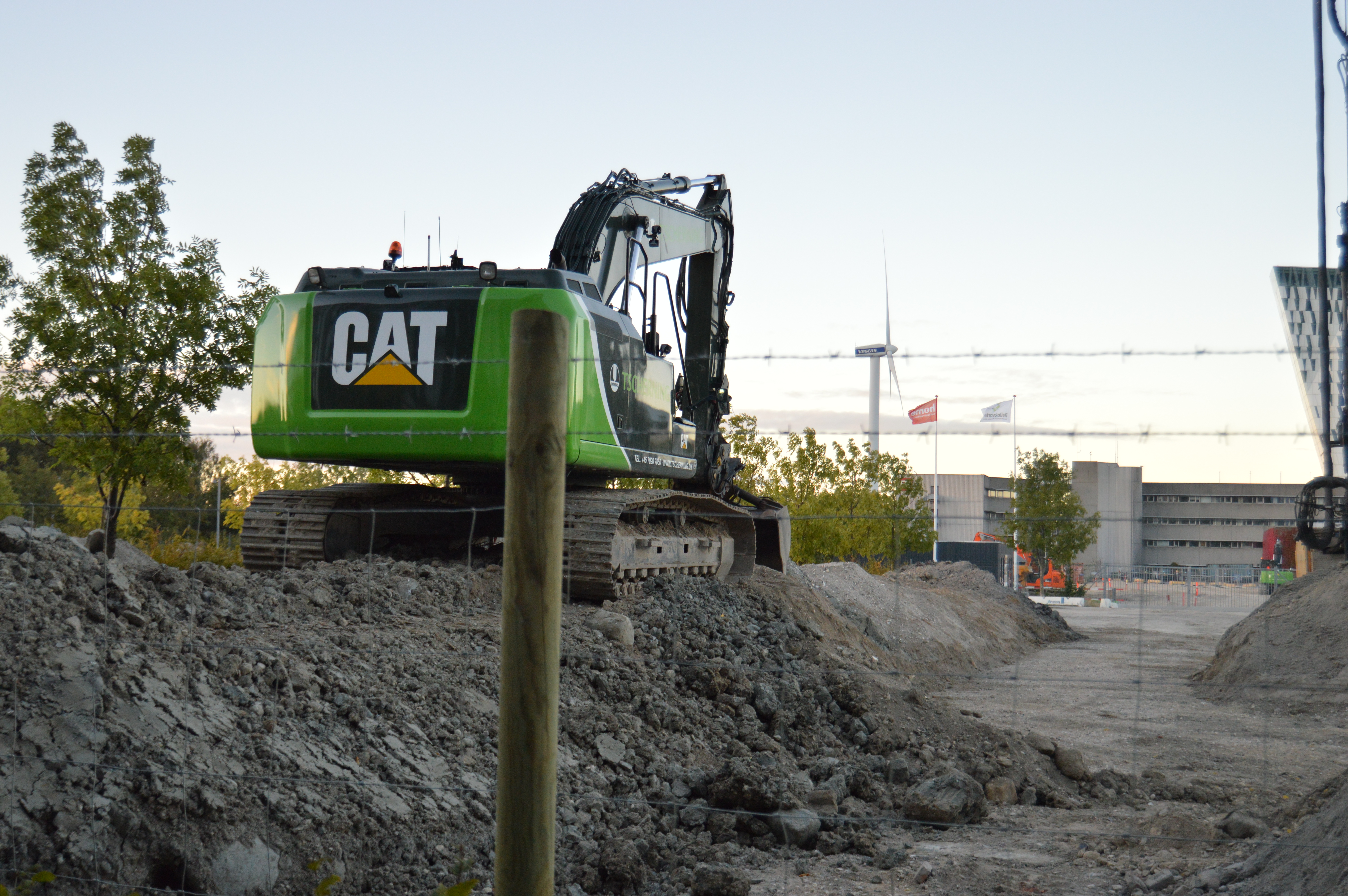 På en byggeplads er en grøn CAT-maskine parkeret. For at øge sikkerheden på pladsen, er der monteret et byggepladshegn